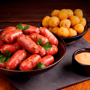 Chorizos cóctel en plato negro con papas criollas doradas y salsa rosada.