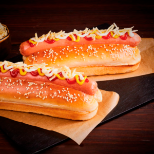 Dos perros calientes estilo americano con salchicha, kétchup, mostaza, mayonesa y queso rallado en pan con ajonjolí.